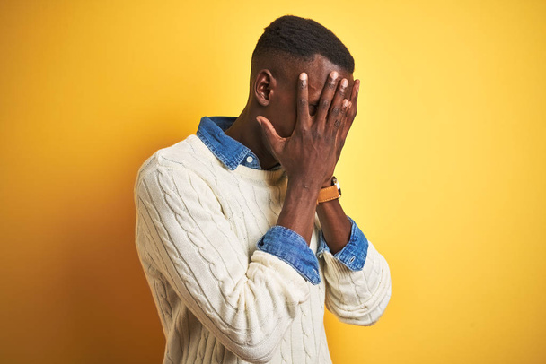 Homem afro-americano vestindo camisa jeans e suéter branco sobre fundo amarelo isolado com expressão triste cobrindo o rosto com as mãos enquanto chora. Conceito de depressão
. - Foto, Imagem