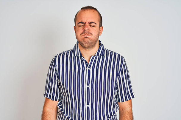 Νεαρός άντρας με μπλε ριγέ πουκάμισο στέκεται πάνω από απομονωμένο λευκό φόντο φουσκώνει μάγουλα με αστείο πρόσωπο. Στόμα φουσκωμένο με αέρα, τρελή έκφραση. - Φωτογραφία, εικόνα