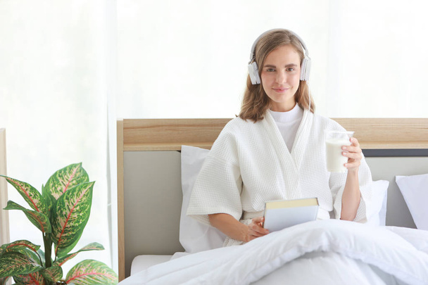 Молодая веселая белая женщина в белой пижаме с наушниками, которая держит стакан молока во время чтения книги перед сном со счастливым и улыбающимся лицом на кровати в белой современной спальне.  - Фото, изображение