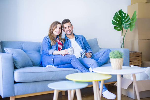 Νεαρό ζευγάρι χαλαρωτικό πίνοντας ένα φλιτζάνι καφέ κάθεται στον καναπέ του νέου σπιτιού με χαρτοκιβώτια γύρω τους, πολύ χαρούμενη μετακίνηση σε ένα νέο διαμέρισμα - Φωτογραφία, εικόνα