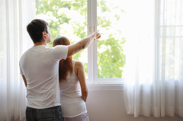 peruutusnäkymä nuori valkoihoinen aviomies syleilee nuoria raskaana olevia aasialaisia vatsa vaimo ja osoittaa jotain ja katso pois ulos ikkunasta
 - Valokuva, kuva