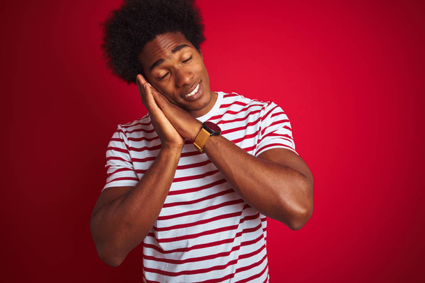 Молодой африканский американец с афроволосами в полосатой футболке на изолированном красном фоне спит уставшим во сне и позирует с руками вместе, улыбаясь с закрытыми глазами
. - Фото, изображение