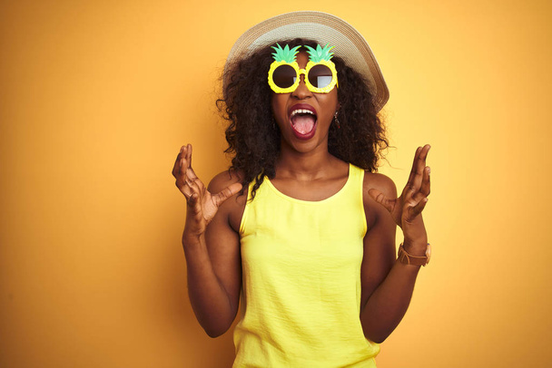 Mulher afro-americana vestindo óculos engraçados de abacaxi sobre fundo amarelo isolado louco e louco gritando e gritando com expressão agressiva e braços levantados. Conceito de frustração
. - Foto, Imagem