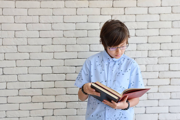cute i inteligentnego małego chłopca w niebieskiej koszulce z okularami gospodarstwa i Reding czerwone książki z zamierzonym twarz i białe tło ścienne - Zdjęcie, obraz