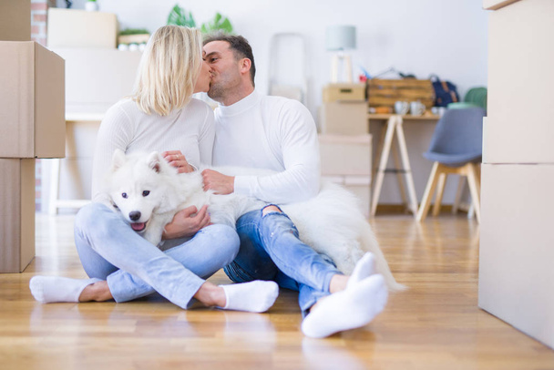 Young beautiful couple with dog kissing sitting on the floor at new home around cardboard boxes - Foto, afbeelding