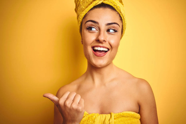 Fiatal gyönyörű nő visel törülközőt zuhany után elszigetelt sárga háttér mutatva, és mutatja hüvelykujjával felfelé az oldalon boldog arc mosolygós - Fotó, kép