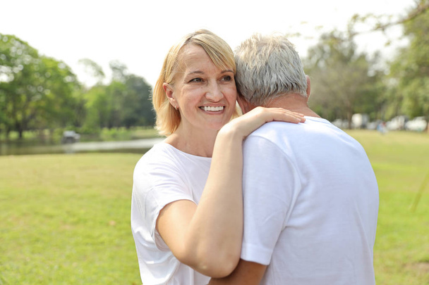 smiling elderly caucasian couple with white shirt and blue jean embracing in park in summer time - Photo, Image