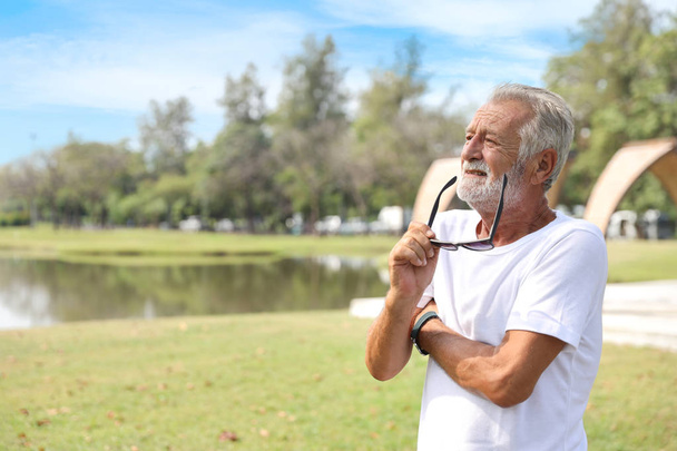 ältere kaukasische Männer, weißer Bart in weißem Hemd und blauer Jeans mit Sonnenbrille ruhen sich aus und leben nach ihrer Pensionierung im Sommer im Park. er schaut oder denkt etwas. - Foto, Bild