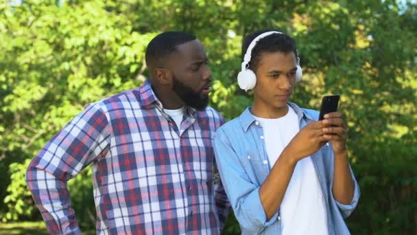 Afroamerikkalainen mies huutaa huomiotta teini poika kuulokkeet älypuhelimella
 - Materiaali, video