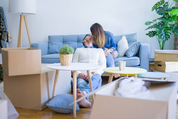 Young beautiful couple sitting on the sofa drinking cup of coffee at new home around cardboard boxes - Photo, Image