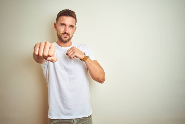 Young handsome man wearing casual white t-shirt over isolated background Punching fist to fight, aggressive and angry attack, threat and violence - Fotografie, Obrázek