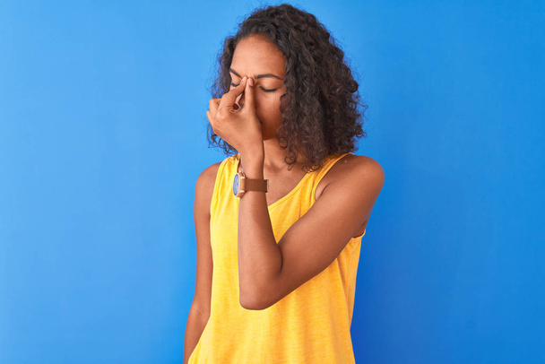 Young brazilian woman wearing yellow t-shirt standing over isolated blue background tired rubbing nose and eyes feeling fatigue and headache. Stress and frustration concept. - Foto, Imagem