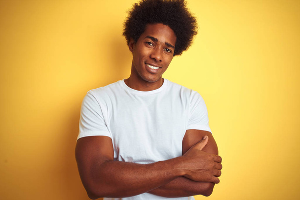 amerikanischer Mann mit Afro-Haaren in weißem T-Shirt vor gelbem Hintergrund, glücklich lächelnd mit verschränkten Armen in die Kamera blickend. positive Person. - Foto, Bild