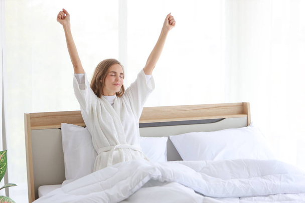 Iloinen ja terve valkoihoinen nuori nainen yllään pyjama herätä aamulla auringonnousun ja istuu ja venyttely sängyssä makuuhuoneessa rentoutumista ja onnellinen kasvot - Valokuva, kuva