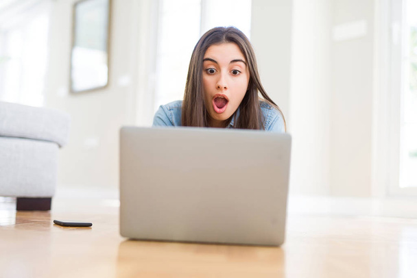 Όμορφη νεαρή γυναίκα που βρίσκονται στο πάτωμα χρησιμοποιώντας το laptop φοβάται σε σοκ με μια έκπληξη πρόσωπο, φοβισμένος και ενθουσιασμένος με την έκφραση του φόβου - Φωτογραφία, εικόνα
