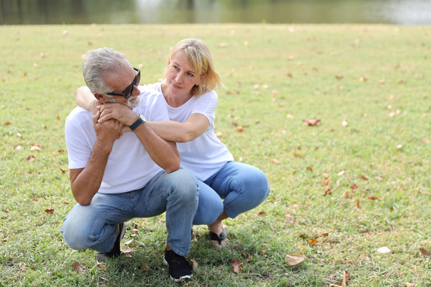 caucasian elderly couple with white shirt, blue jean and sun glasses sitting and embracing in park during summer time on wedding anniversary day - Photo, Image
