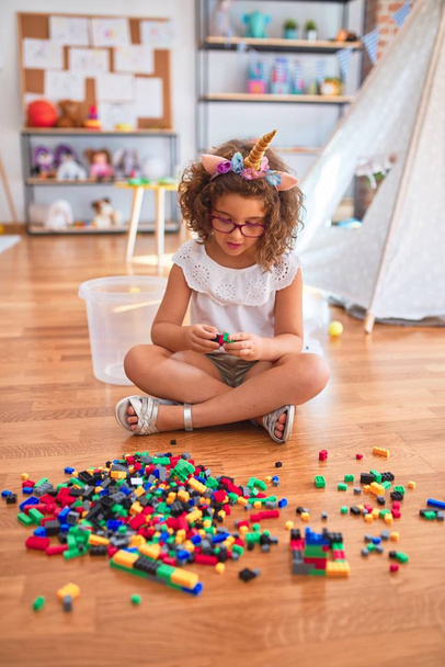schönes Kleinkind mit Brille und Einhorn-Diadem, das im Kindergarten auf dem Boden sitzt und mit Bausteinen spielt - Foto, Bild