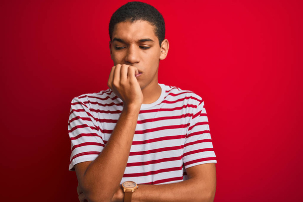 Jonge knappe Arabier man draagt gestreept t-shirt over geïsoleerde rode achtergrond kijken gestrest en nerveus met handen op de mond bijtende nagels. Angst probleem. - Foto, afbeelding