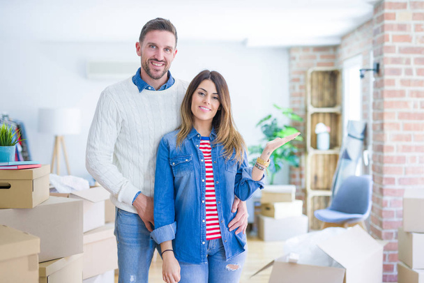 Young beautiful couple standing at new home around cardboard boxes smiling cheerful presenting and pointing with palm of hand looking at the camera. - Foto, immagini