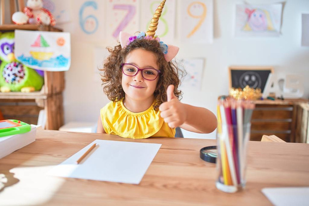 Hermoso niño pequeño con gafas y diadema de unicornio sentado en el escritorio en el jardín de infantes haciendo un gesto feliz pulgares hacia arriba con la mano. Aprobar la expresión mirando a la cámara con éxito
. - Foto, imagen