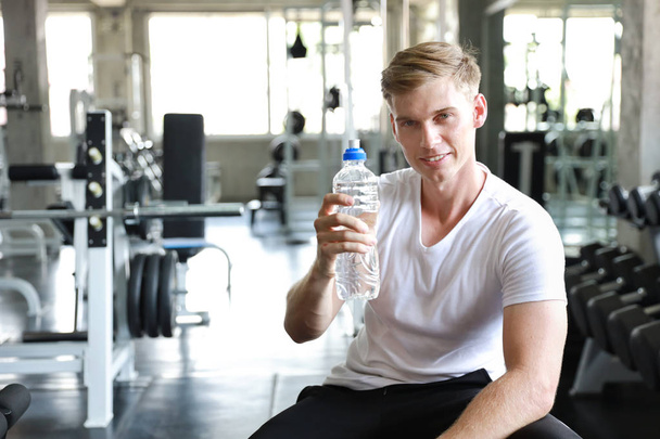 Przystojny kaukaski mężczyzna trzyma i picie czystej butelki wody w fitness z uśmiechniętą twarzą - Zdjęcie, obraz