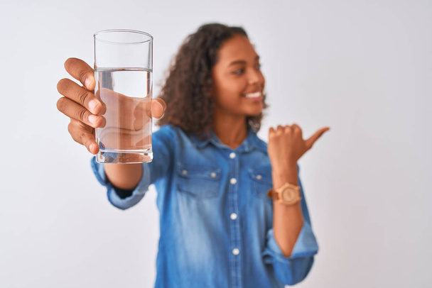 Mladá brazilská žena drží sklenici vody stojí nad izolovaným bílým pozadím ukazuje palcem nahoru na stranu se šťastnou tváří usmívá - Fotografie, Obrázek