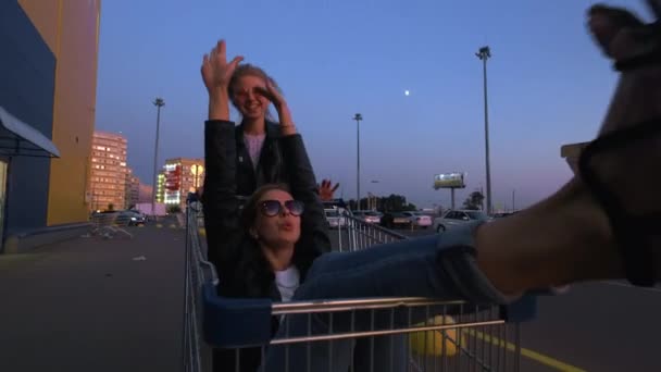 ストリートウェアを着た2人のミレニアル世代のガールフレンドは、日没時にスーパーマーケットの駐車場で楽しんでいます。ショッピングカートに乗って、自由を楽しむ - 映像、動画