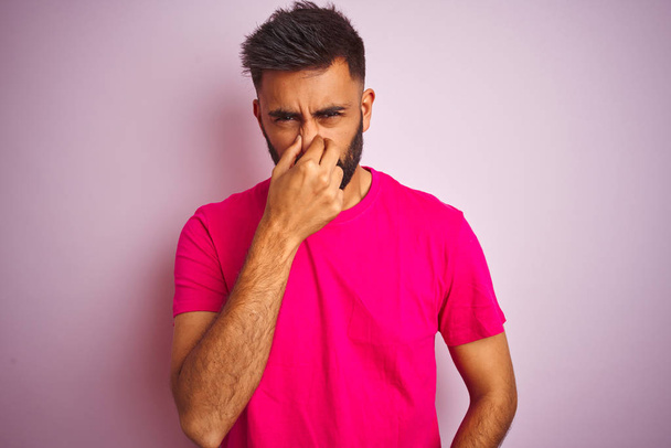 junger indischer Mann mit T-Shirt, der vor isoliertem rosa Hintergrund steht und etwas Stinkendes und ekelhaften, unerträglichen Geruch riecht und mit den Fingern auf der Nase den Atem anhält. Konzept für schlechte Gerüche. - Foto, Bild