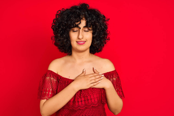 Mujer árabe joven con el pelo rizado con vestido casual sobre fondo rojo aislado sonriendo con las manos en el pecho con los ojos cerrados y gesto agradecido en la cara. Concepto de salud
. - Foto, Imagen