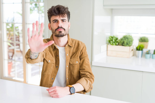 Young man wearing casual jacket sitting on white table doing stop sing with palm of the hand. Warning expression with negative and serious gesture on the face. - 写真・画像