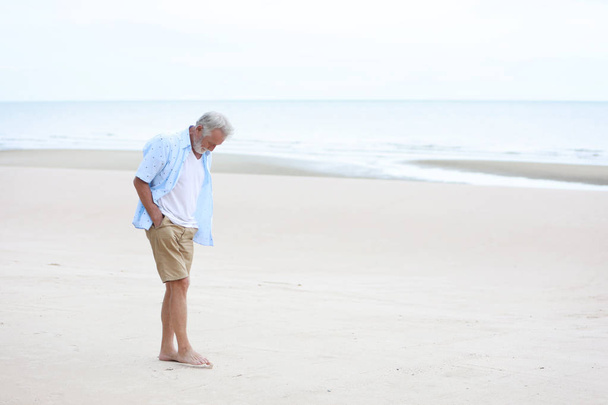 full shot image of older caucasian men, white beard and white hair in white and blue shirt is resting and standing on sandy beach after retirement in resort during the summer with right copy space - 写真・画像