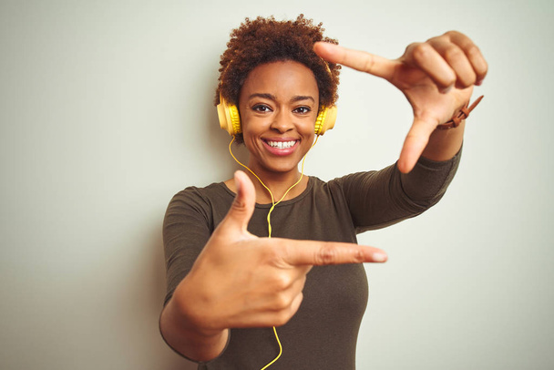 Африканська американка, одягнена в навушники, слухаючи музику через ізольований фон, посміхається, роблячи рамку руками і пальцями з щасливим обличчям. Творчість і фотографія. - Фото, зображення