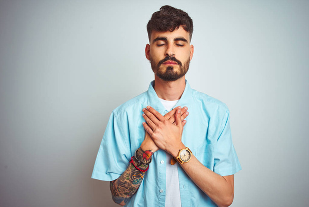 Young man with tattoo wearing blue shirt standing over isolated white background smiling with hands on chest with closed eyes and grateful gesture on face. Health concept. - Photo, Image