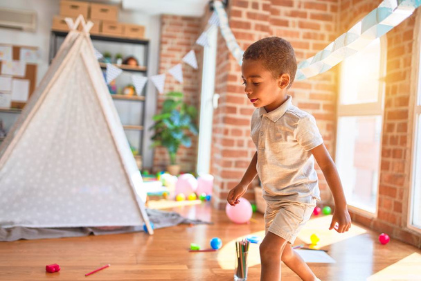 Beautiful african american toddler standing and smiling around lots of toys at kindergarten - 写真・画像
