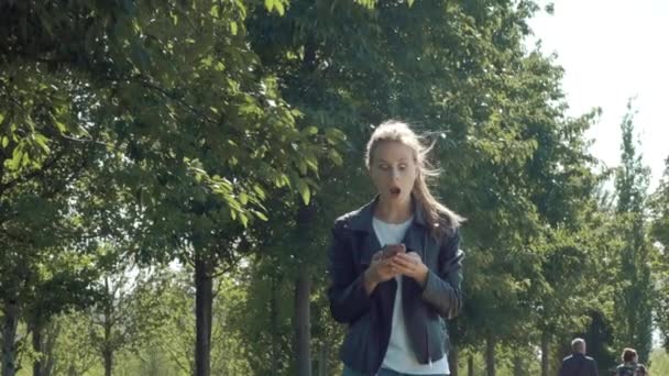 Young beautiful girl is walking in the park with a smartphone. Reads the message and is very happy. Dancing and bouncing with happiness and joy. - Séquence, vidéo