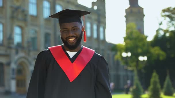 Zadowolony czarny mężczyzna w sukni dyplomowej uśmiechnięty aparat fotograficzny na tle uczelni - Materiał filmowy, wideo