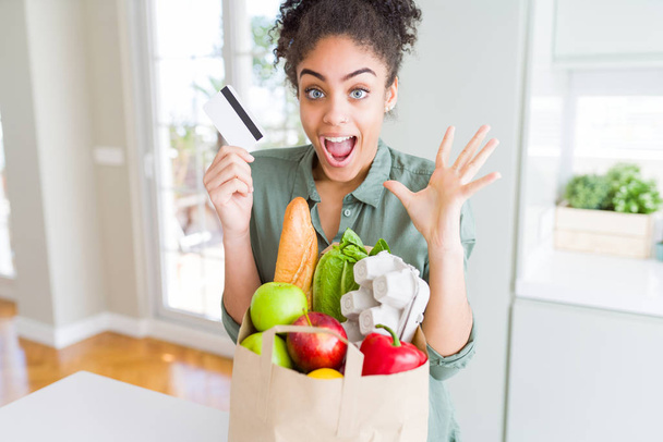 Young african american girl holding paper bag of groceries and credit card as payment very happy and excited, winner expression celebrating victory screaming with big smile and raised hands - Fotografie, Obrázek