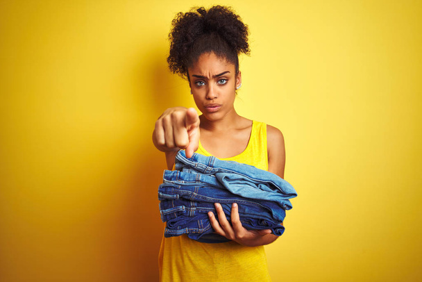 Young african american woman holding stack of jeans over isolated yellow background pointing with finger to the camera and to you, hand sign, positive and confident gesture from the front - Photo, Image
