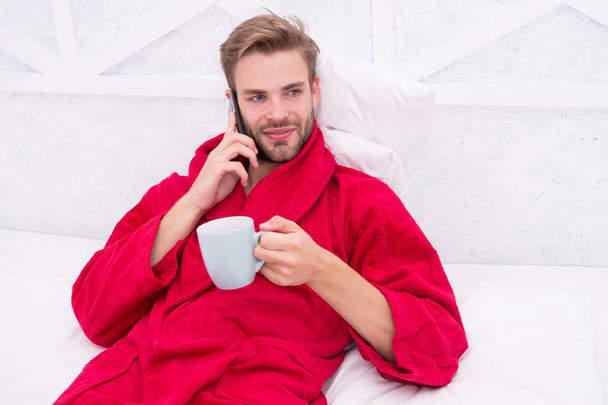 Coffee and technology have gone hand in hand. Young man using mobile technology in bed. Handsome guy talking on phone and drinking coffe at home. Modern life and new technology. Technology concept - Photo, image