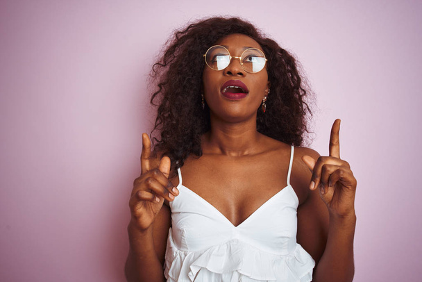 Mujer afroamericana joven con gafas de pie sobre fondo rosa aislado asombrado y sorprendido mirando hacia arriba y señalando con los dedos y los brazos levantados
. - Foto, imagen