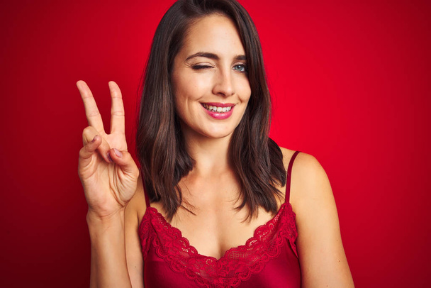 Νέα όμορφη γυναίκα φορώντας σέξι εσώρουχα πάνω από το κόκκινο απομονωμένο φόντο χαμογελά με χαρούμενο πρόσωπο που κλείνει το μάτι στην κάμερα κάνει το σημάδι της νίκης. Νούμερο δύο.. - Φωτογραφία, εικόνα