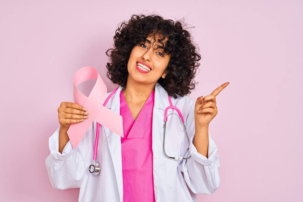 Young arab doctor woman with curly hair holding cancer ribbon over isolated pink background very happy pointing with hand and finger to the side - Photo, Image