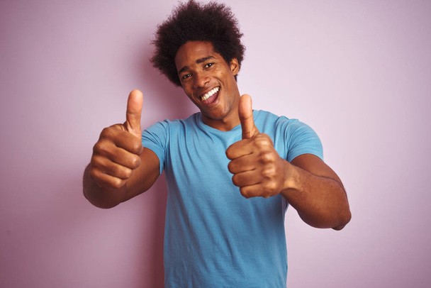Afrikanisch-amerikanischer Mann mit Afro-Haaren und blauem T-Shirt, der vor isoliertem rosa Hintergrund steht und eine positive Geste mit der Hand macht, die Daumen hoch lächelnd und glücklich über den Erfolg. Siegergeste. - Foto, Bild