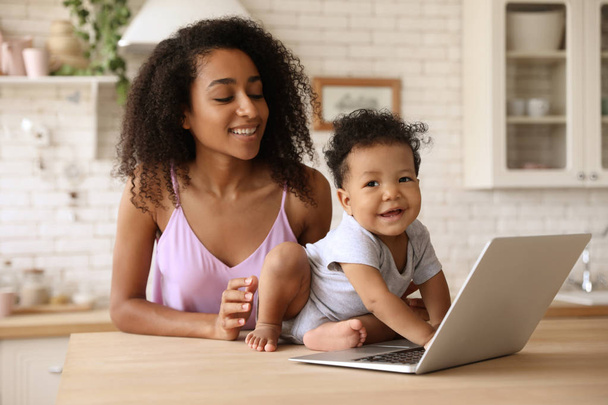 Mulher afro-americana e seu bebê com laptop na cozinha. Felicidade da maternidade
 - Foto, Imagem