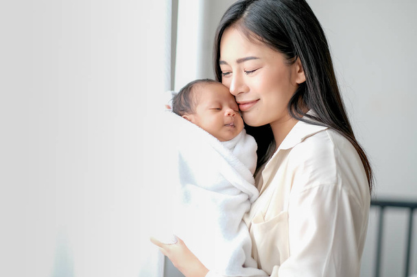 Wit overhemd Aziatische moeder is zoenen haar pasgeboren baby in de slaapkamer voor glasramen met wit gordijn om liefde en familie bonding tonen. - Foto, afbeelding
