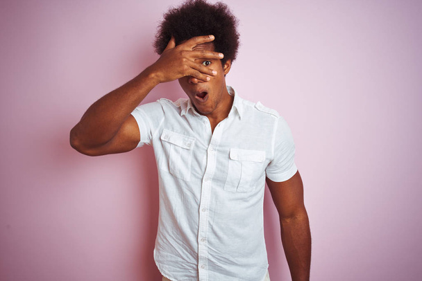Fiatal amerikai férfi afro haj viselt fehér ing felett áll elszigetelt rózsaszín háttér kukucskál a sokk fedő arc és a szemek kézzel, néz keresztül ujjak zavarba ejtő kifejezés. - Fotó, kép
