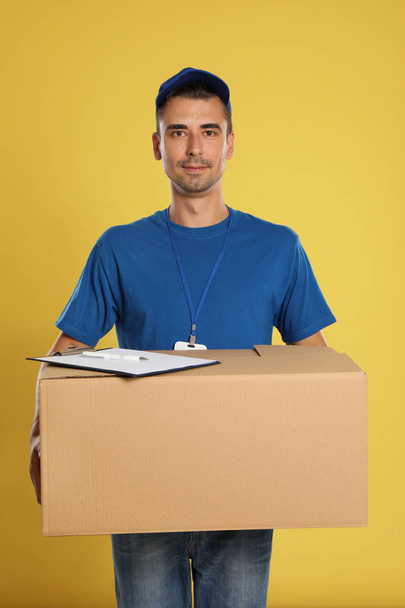 Happy young courier with cardboard box and clipboard on yellow background - Valokuva, kuva