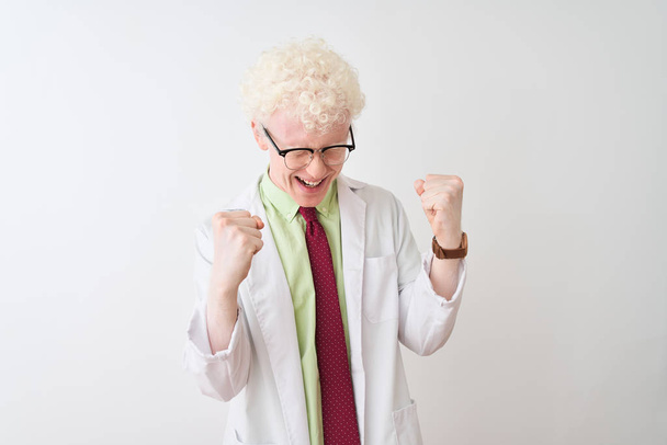 Νέοι Albino ξανθιά επιστήμονας άνθρωπος φορώντας παλτό και γυαλιά πάνω από απομονωμένο λευκό φόντο πολύ χαρούμενος και ενθουσιασμένος κάνει χειρονομία νικητής με τα χέρια έθεσε, χαμογελώντας και ουρλιάζοντας για την επιτυχία. Έννοια εορτασμού. - Φωτογραφία, εικόνα