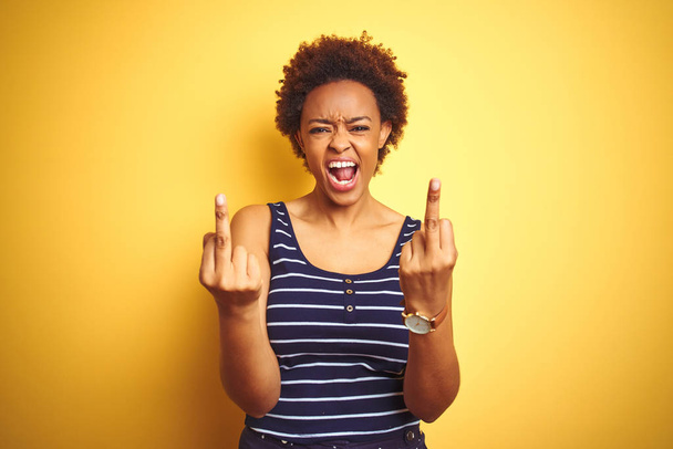 Beauitul african american woman wearing summer t-shirt over isolated yellow background Showing middle finger doing fuck you bad expression, provocation and rude attitude. Screaming excited - Photo, Image