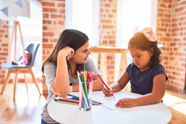 Güzel öğretmen ve küçük kız anaokulunda renkli kalemlerle resim çiziyor. - Fotoğraf, Görsel